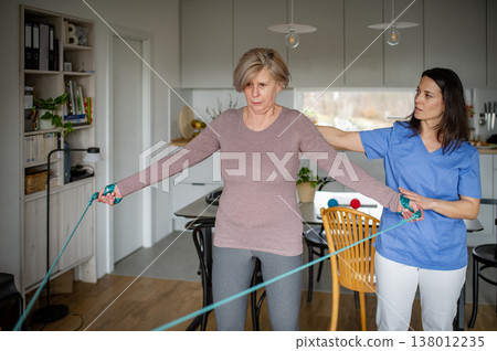 Physiotherapist guiding senior woman during resistance band exercise. 138012235
