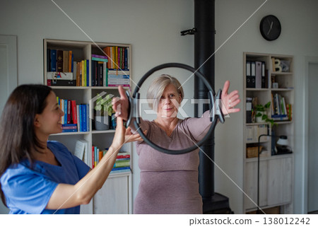 Senior woman performing physiotherapy exercise with ring under physiotherapist supervision at home. Senior woman performing physiotherapy exercise with ring under physiotherapist supervision at home. 138012242
