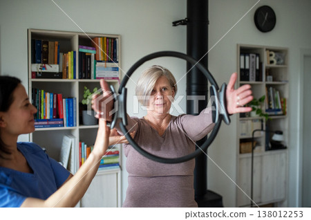 Senior woman performing physiotherapy exercise with ring under physiotherapist supervision at home. Senior woman performing physiotherapy exercise with ring under physiotherapist supervision at home. 138012253