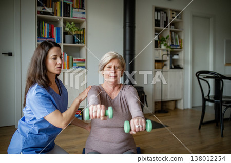 Physiotherapist assisting senior woman with dumbbell exercise, home rehabilitation. 138012254