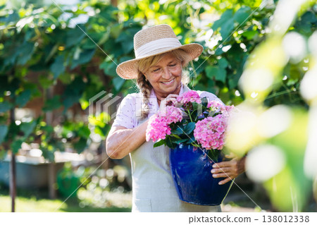 Smiling senior woman holding pot of pink flowers in garden. Smiling senior woman holding pot of pink flowers in garden. 138012338