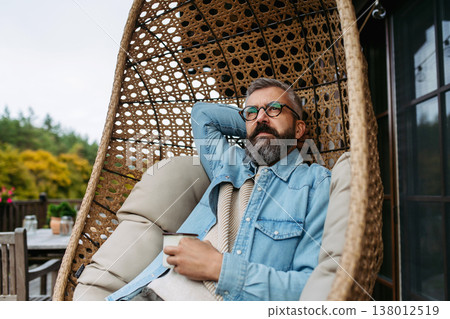 Man is enjoying cup of warm tea, coffee, having relaxing moment at home, sitting in hanging chair on patio. 138012519