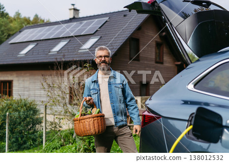 Man with basket with groceries charging eletric car. House with solar panels on roof behind. 138012532