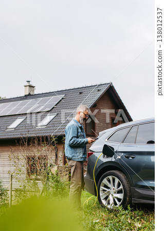 Charging electric car at home, using app. Solar panels on roof behind him. 138012537