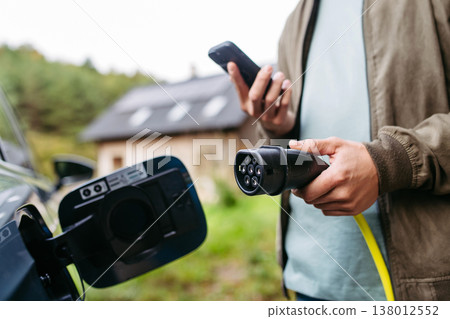 Man charging electric car in front of his house, plugging the charger into the charging port. House with solar panel system on roof behind him. 138012552