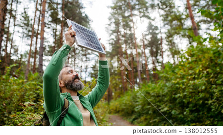 Handsome man standing in the middle of forest, holding small solar panel above his head. Handsome man standing in the middle of forest, holding small solar panel above his head. 138012555