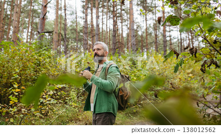 Handsome man watching wildlife with binoculars, enjoying peaceful atmosphere of forest, forestbathing Handsome man watching wildlife with binoculars, enjoying peaceful atmosphere of forest, forestbathing 138012562