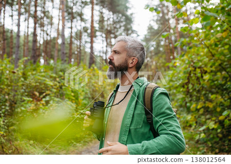 Handsome man watching wildlife with binoculars, enjoying peaceful atmosphere of forest, forestbathing Handsome man watching wildlife with binoculars, enjoying peaceful atmosphere of forest, forestbathing 138012564