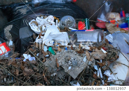 Household waste, glass bottles, and cigarette butts piled on dry leaves inside a dumpster. Close-up view highlighting Household waste, glass bottles, and cigarette butts piled on dry leaves inside a dumpster. Close-up view highlighting 138012937