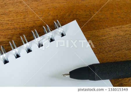 Blank spiral notebook on a wooden desk with a black pen at the lower edge, with customizable space on notebook for text or idea 138015349