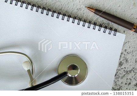 White spiral notebook with a doctor stethoscope and pen arranged on a neutral background, with customizable space on notebook for text or ideas 138015351