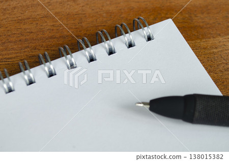 Blank spiral notebook placed on wooden surface with black pen entering frame, With customizable space on notebook for text or ideas 138015382