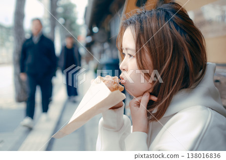 一位二十多歲的女子一邊在旅遊景點漫步,一邊享用新鮮出爐的麵包。 一位二十多歲的女子一邊在旅遊景點漫步,一邊享用新鮮出爐的麵包。 138016836
