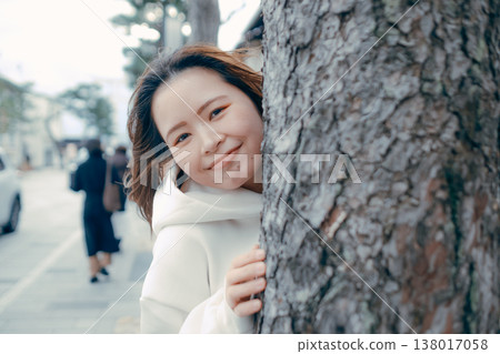 A woman in her 20s peeking out from behind a tree in the city. A woman in her 20s peeking out from behind a tree in the city. 138017058