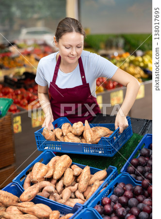 Smiling saleswoman lying out boxes of sweet potatoes in farmers market Smiling saleswoman lying out boxes of sweet potatoes in farmers market 138017695