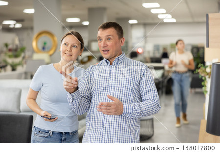 In company accompanied by husband woman walks through furniture store with mobile phone in hands. In company accompanied by husband woman walks through furniture store with mobile phone in hands. 138017800