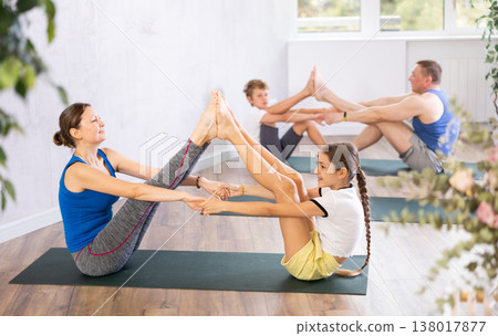 Middle-aged mom and dad and their kids practicing yoga partner in pairs on mats during family yoga class in wellness studio 138017877