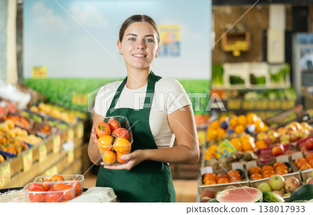 Positive saleswoman in apron offering persimmon on supermarket 138017933