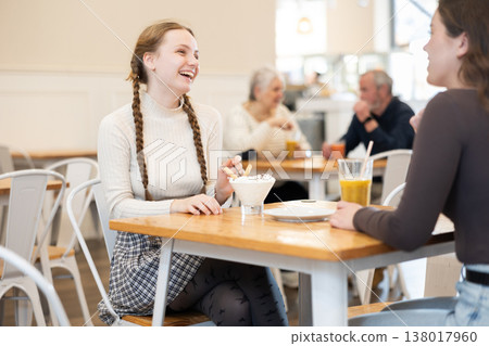 Two positive girls chatting over juice and eat ice cream while sitting at small table in cafe Two positive girls chatting over juice and eat ice cream while sitting at small table in cafe 138017960