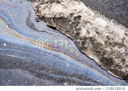Oil slick creating iridescent rainbow pattern on wet asphalt road next to dirty melting snow 138018018