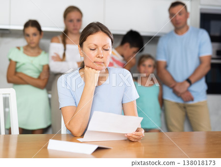 Upset mum is sitting in kitchen, reading letter. Blurred worried family in background 138018173