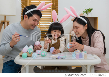 father, mother and daughter wearing bunny ears painting and decorating egg  together at home on easter day. happy family holiday activity concept 138018262