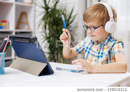Young boy with headphones studying at desk with tablet and notebook Young boy with headphones studying at desk with tablet and notebook 138018312