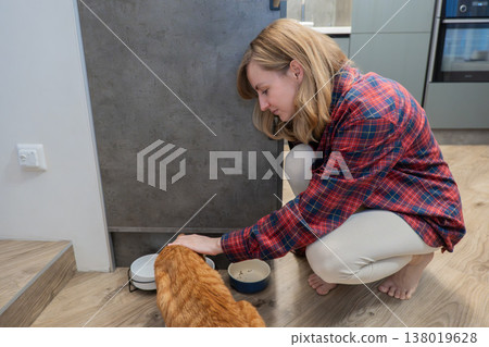 Caucasian lady gently petting warm orange kitten beside dishes in evening lighting, Woman of Caucasian appearance tenderly strokes vibrant orange cat near bowls in cozy evening kitchen scene Caucasian lady gently petting warm orange kitten beside dishes in evening lighting, Woman of Caucasian appearance tenderly strokes vibrant orange cat near bowls in cozy evening kitchen scene 138019628