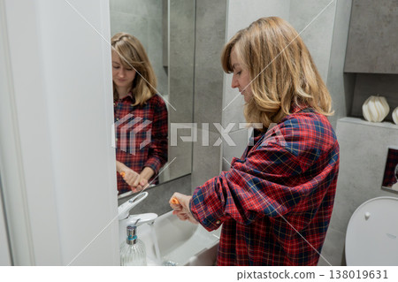 Woman cleaning bathroom sink and mirror with cloth, focused on wiping dust from faucet and vanity, plaid shirt, reflective mirror showing profile, tidy contemporary bathroom, soft natural light, calm 138019631