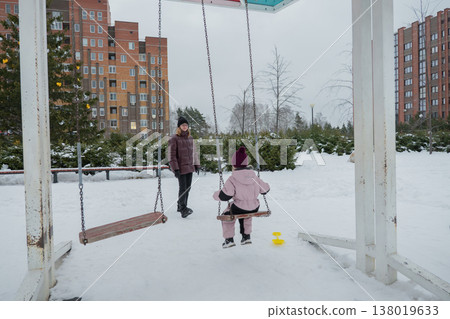 snowy playground caucasian woman pushing toddler in winter jackets and mittens under metal swing frame near apartment buildings overcast sky yellow toy at feet intimate urban caregiving moment showing 138019633