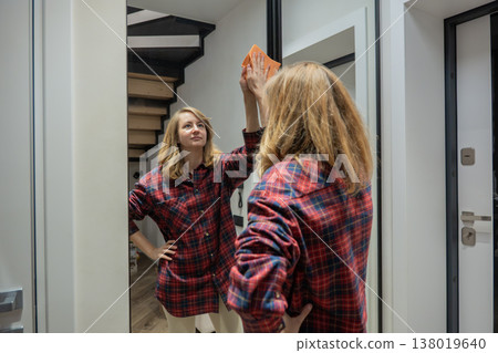 caucasian woman dusting mirror in hallway with plaid shirt and soft smile, wiping reflection and wood trim, bright modern staircase visible behind, casual morning routine showing tidy domestic upkeep caucasian woman dusting mirror in hallway with plaid shirt and soft smile, wiping reflection and wood trim, bright modern staircase visible behind, casual morning routine showing tidy domestic upkeep 138019640