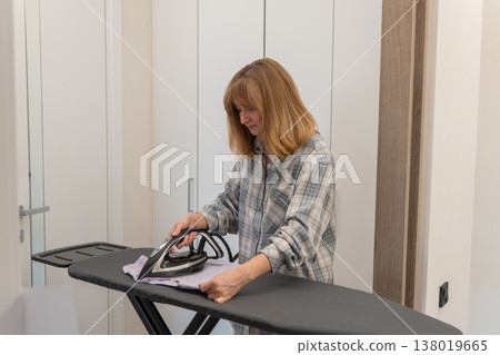 Woman ironing plaid shirt on board in bright hallway, focused on smoothing fabric with steam iron, casual loungewear and tidy interior doors in background, quiet domestic chore and relaxed lifestyle Woman ironing plaid shirt on board in bright hallway, focused on smoothing fabric with steam iron, casual loungewear and tidy interior doors in background, quiet domestic chore and relaxed lifestyle 138019665