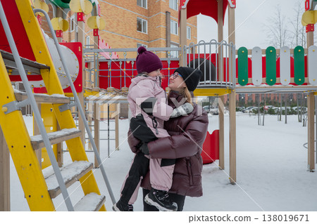 Caucasian caregiver comforting daughter by ladder at snowcovered playground, attentive adult holds child steady and reassures with gentle hug, bundled winter layers and knit hats, residential Caucasian caregiver comforting daughter by ladder at snowcovered playground, attentive adult holds child steady and reassures with gentle hug, bundled winter layers and knit hats, residential 138019671