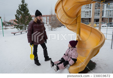 caucasian mother supervising daughter on snowy playground slide, holding yellow shovel, bundled puffer coats, knit hats, boots gentle smile, watching play, visible breath, apartment buildings caucasian mother supervising daughter on snowy playground slide, holding yellow shovel, bundled puffer coats, knit hats, boots gentle smile, watching play, visible breath, apartment buildings 138019672