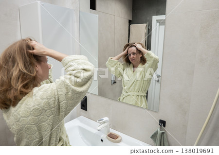 Caucasian woman at mirror checking expression while running hands through hair in bathrobe, serious concentrated look, modern bathroom sink and shower visible, preparing for audition with tense 138019691