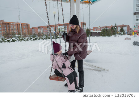 Caucasian caregiver gently pushing little girl on snowy swing while monitoring safety, maroon puffer, pink snowsuit and knit hat, playground chain motion, warm connection and mindful parenting Caucasian caregiver gently pushing little girl on snowy swing while monitoring safety, maroon puffer, pink snowsuit and knit hat, playground chain motion, warm connection and mindful parenting 138019703