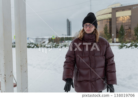 Caucasian woman walking through snowy park area after play, maroon puffer and black beanie, thoughtful expression, footprints trailing in fresh snow, apartment blocks and overcast sky, reflective 138019706