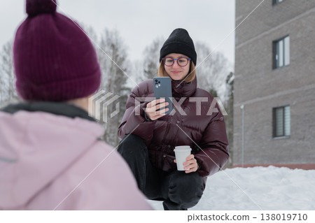 Woman checking phone while holding coffee, closeup portrait in snowy courtyard, black beanie and glasses, maroon puffer jacket, relaxed expression with slight smile, smartphone screen visible, urban 138019710