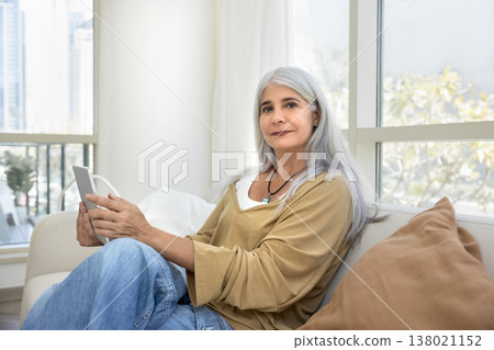 Mature female sits on couch with tablet, looking at camera 138021152