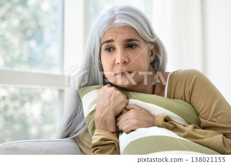 Mature woman hugging pillow seated on sofa feels defenseless Mature woman hugging pillow seated on sofa feels defenseless 138021155