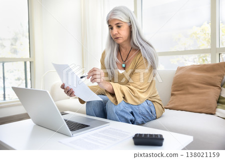 Serious middle aged woman reviewing documents seated on sofa 138021159