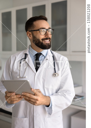 Cheerful smart young male doctor holding touchscreen tablet looking away Cheerful smart young male doctor holding touchscreen tablet looking away 138021208