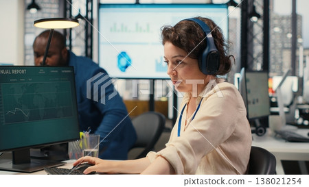 Female executive consultant listening to songs on headphones at the workstation, managing administrative tasks to ensure company productivity and efficiency. Woman reviews data. Camera B. 138021254