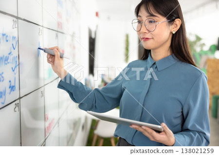 Positive young Asian manager woman writing notes on office whiteboard Positive young Asian manager woman writing notes on office whiteboard 138021259