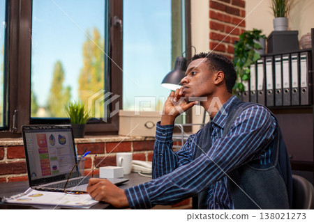 Tired male analyst holding his chin at desk and staring outside the window in brick wall workspace. Manager leans head on his hand, feeling exhausted by business analytics research at startup office. Tired male analyst holding his chin at desk and staring outside the window in brick wall workspace. Manager leans head on his hand, feeling exhausted by business analytics research at startup office. 138021273