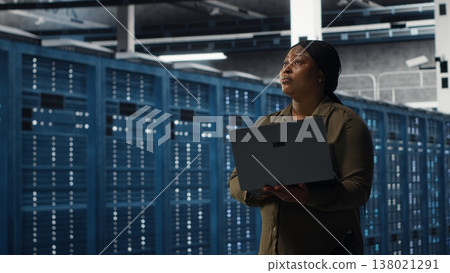 Smiling engineer walking in data center with laptop, checking hardware for system stability. Upbeat employee overseeing server farm with notebook, configuring infrastructure, camera B Smiling engineer walking in data center with laptop, checking hardware for system stability. Upbeat employee overseeing server farm with notebook, configuring infrastructure, camera B 138021291