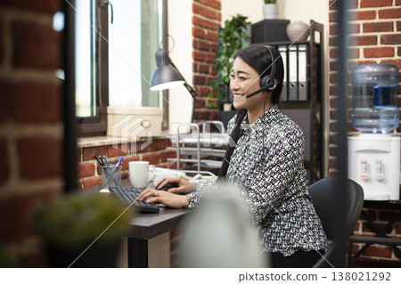 Female employee uses desktop pc, typing on keyboard and engaging in a lively conversation through wired headphones in modern workplace. Smiling woman seated at office desk, using computer and headset. Female employee uses desktop pc, typing on keyboard and engaging in a lively conversation through wired headphones in modern workplace. Smiling woman seated at office desk, using computer and headset. 138021292