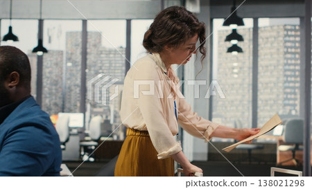 Workplace support employee moves a delivery cart stacked with folders and contracts, helping african american administrator bringing stack of archived documents for accounting tasks. Camera B. 138021298