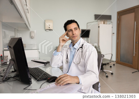 Pensive male doctor in white lab coat sitting at desk Pensive male doctor in white lab coat sitting at desk 138021303