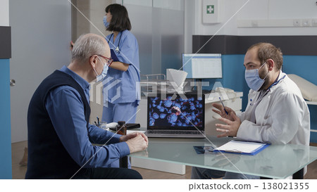 Physician explaining coronavirus spreading with illustration on laptop, giving medical advice to elder man at checkup visit. Medic and patient looking at virus animation on display. 138021355